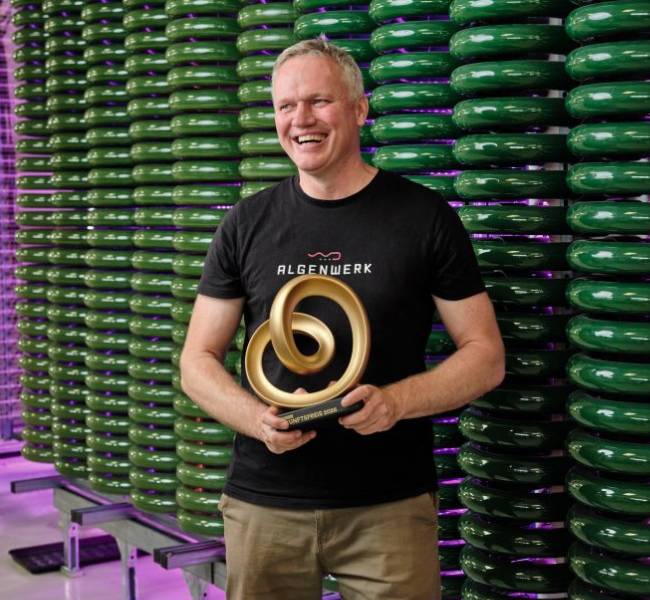 Zu sehen ist Algenwerk Gründer Gunnar Mühlstädt mit dem Zukunftspreis in der Hand vor einer Algenanlage