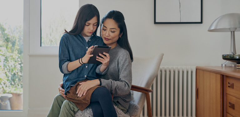 Ein Mädchen sitzt mit ihrem Tablet in der Hand auf dem Schoß ihrer Mutter und erklärt ihr etwas. Die Mutter hört gespannt zu. Beide befinden sich in einem hellen und modernen Raum.