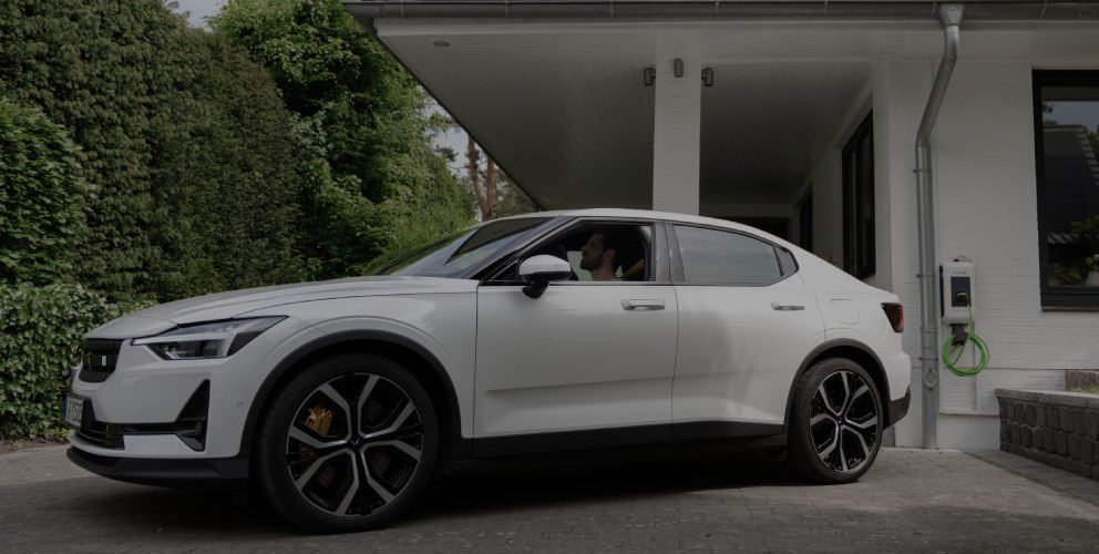 Hinweis: 85 € THG-Prämie für 2025 sichern! Ein Mann sitzt in einem Elektroauto vor einem Einfamilienhaus.
