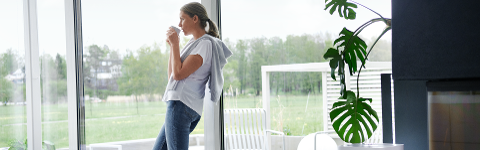 Eine Frau steht an einer Glasfront mit Blick auf einen ordentlichen Garten. Sie hält eine Tasse in beiden Händen und trinkt daraus. Im Vordergrund ist eine Zimmerpflanze zu sehen.