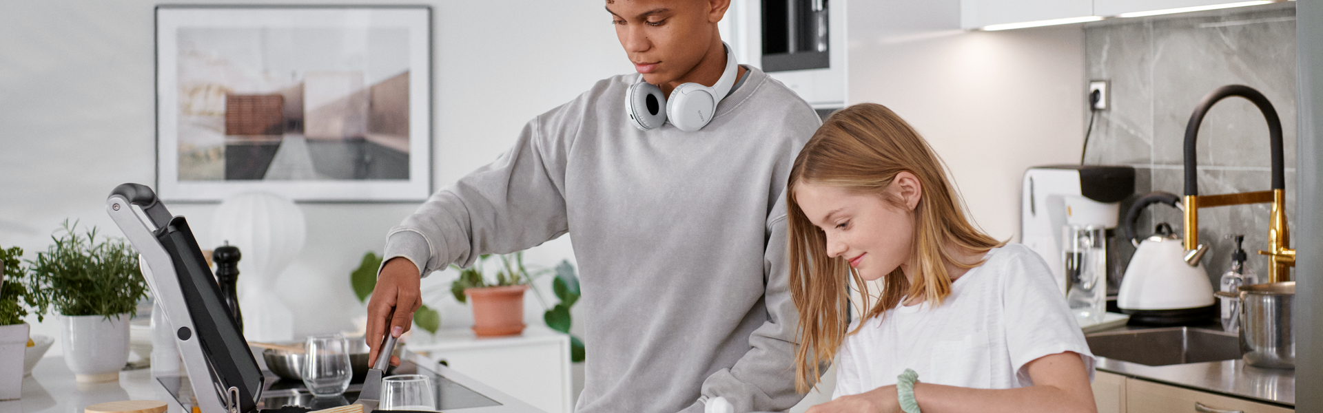 Zwei Teenager stehen in einer modernen und hellen Küche. Der Junge bereitet etwas vor und hat kabellose Kopfhörer um seinen Hals. Das Mädchen steht freudig daneben und hilft ihm.
