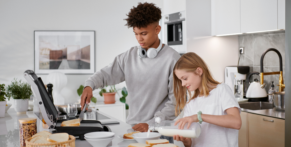 Zwei Teenager stehen in einer modernen und hellen Küche. Der Junge bereitet etwas vor und hat kabellose Kopfhörer um seinen Hals. Das Mädchen steht freudig daneben und hilft ihm.