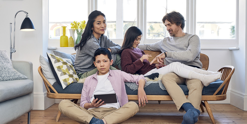 Eine Familie, bestehend aus einem Sohn, einer Tochter, einem Vater und einer Mutter, sitzt in einem hellen und modernen Wohnzimmer auf einer gemütlichen Bank, die einem Sofa ähnelt.