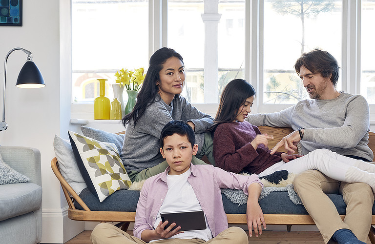Eine Familie, bestehend aus einem Sohn, einer Tochter, einem Vater und einer Mutter, sitzt in einem hellen und modernen Wohnzimmer auf einer gemütlichen Bank, die einem Sofa ähnelt.