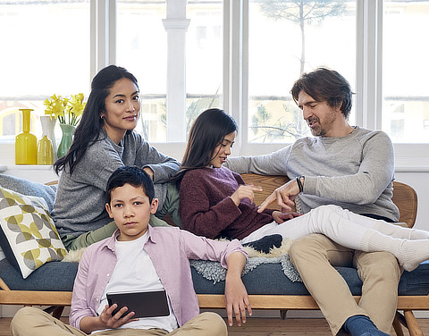 Eine Familie, bestehend aus einem Sohn, einer Tochter, einem Vater und einer Mutter, sitzt in einem hellen und modernen Wohnzimmer auf einer gemütlichen Bank, die einem Sofa ähnelt.