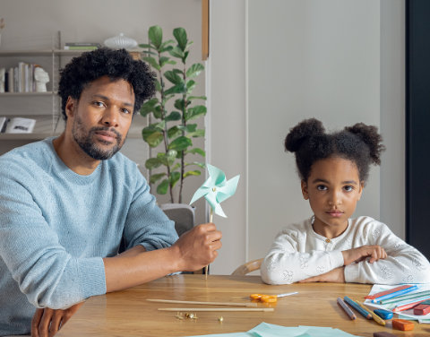 Ein Vater sitzt mit seiner Tochter in einem modernen Wohnzimmer und hält ein kleines türkisenes Windrad in der Hand.