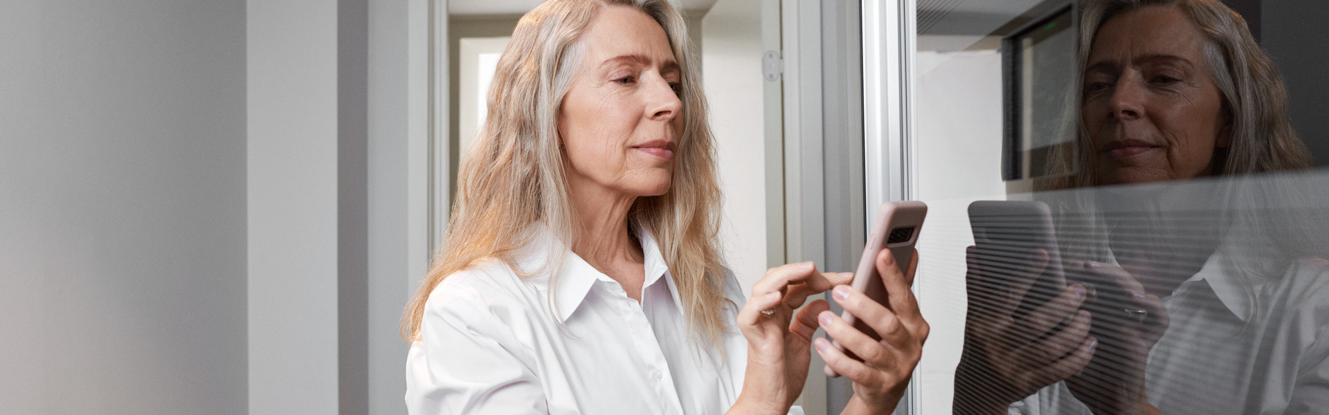 Frau hält ihr Smartphone in der Hand und wählt mit ihrem Zeigefinger etwas aus. Sie befindet sich in einem hellen weißen Raum am Fenster.
