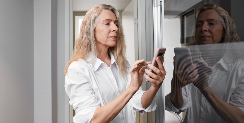 Frau hält ihr Smartphone in der Hand und wählt mit ihrem Zeigefinger etwas aus. Sie befindet sich in einem hellen weißen Raum am Fenster.