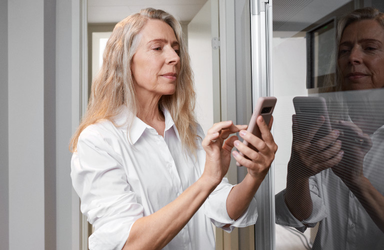 Frau hält ihr Smartphone in der Hand und wählt mit ihrem Zeigefinger etwas aus. Sie befindet sich in einem hellen weißen Raum am Fenster.