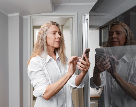 Frau hält ihr Smartphone in der Hand und wählt mit ihrem Zeigefinger etwas aus. Sie befindet sich in einem hellen weißen Raum am Fenster.