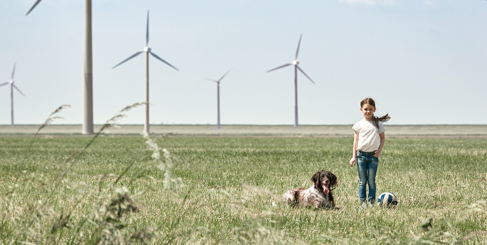 Ein Mädchen steht auf einer sommerlich grünen Wiese. Neben ihr sitzt ein Hund und ein Volleyball im Gras . Im Hintergrund stehen mehrere Windräder.