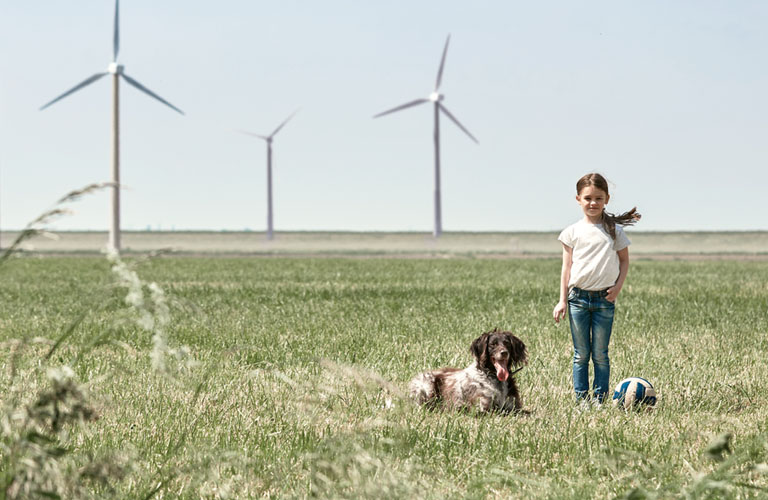 Ein Mädchen steht auf einer sommerlich grünen Wiese. Neben ihr sitzt ein Hund und ein Volleyball im Gras . Im Hintergrund stehen mehrere Windräder.