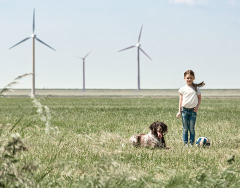 Ein Mädchen steht auf einer sommerlich grünen Wiese. Neben ihr sitzt ein Hund und ein Volleyball im Gras . Im Hintergrund stehen mehrere Windräder.