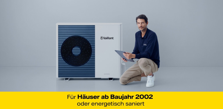 Ein casual gekleideter Mann kniet mit einem Bein neben einer modernen Wärmepumpe. Er hält ein Tablet in der Hand. Der Hintergrund ist schlicht und hell.