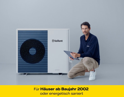 Ein casual gekleideter Mann kniet mit einem Bein neben einer modernen Wärmepumpe. Er hält ein Tablet in der Hand. Der Hintergrund ist schlicht und hell.