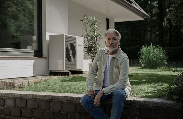 Ein Mann mit einem grauen Bart sitzt vor seinem Haus im Garten. Im Hintergrund ist eine moderne Wärmepumpe.