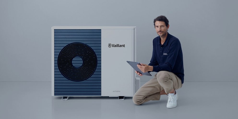 Ein casual gekleideter Mann kniet mit einem Bein neben einer modernen Wärmepumpe. Er hält ein Tablet in der Hand. Der Hintergrund ist schlicht und hell.