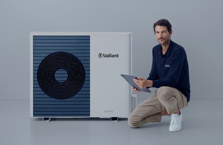 Ein casual gekleideter Mann kniet mit einem Bein neben einer modernen Wärmepumpe. Er hält ein Tablet in der Hand. Der Hintergrund ist schlicht und hell.