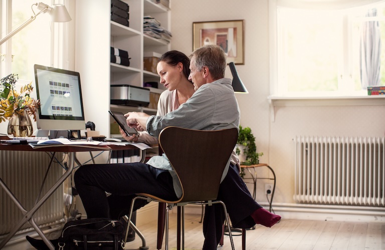 Mann und Frau sitzen in einem modernen, hellen Arbeitszimmer. Die Frau hält ein Tablet in der Hand und der Mann erklärt ihr etwas darauf. Sie hört gespannt zu.