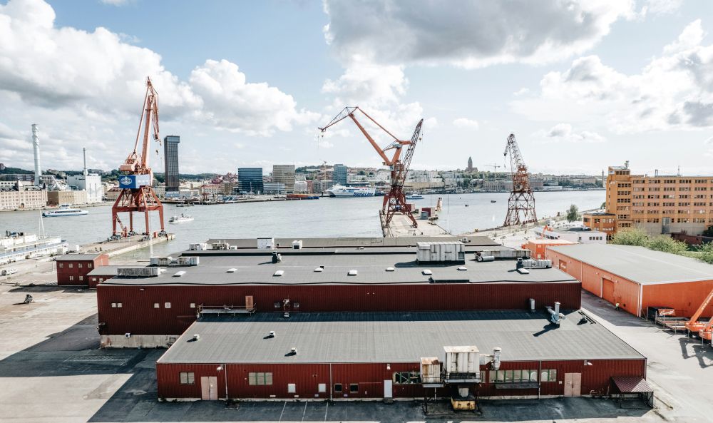 Industriehafen mit Lagerhallen und Hafenkränen, im Hintergrund eine Stadtsilhouette am Wasser.