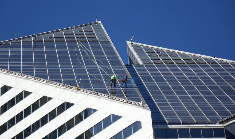 Zwei Arbeiter installieren Solarpaneele auf dem Dach eines modernen Gewerbegebäudes vor blauem Himmel.