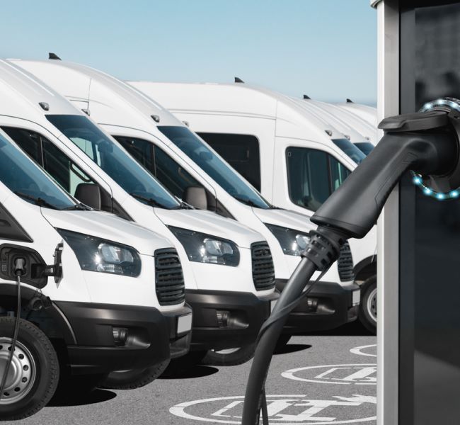 Dieses Bild zeigt elektrische Lieferwagen und eine Ladestation davor. Es symbolisiert den Umstieg auf E-Mobilität im Kontext von Netto-Null-Emissionen (Net Zero) für Unternehmen