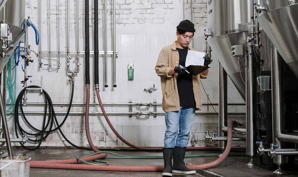 Foto von einem Mann mittleren Alters, der eine blaue Schürze trägt, vor Rohren steht und mit in die Seite gestützten Händen direkt in die Kamera schaut