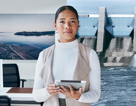 Frau hält Tablet in der Hand. Der Hintergrund zeigt ein Büroraum und eine Wasserkraftanlage.
