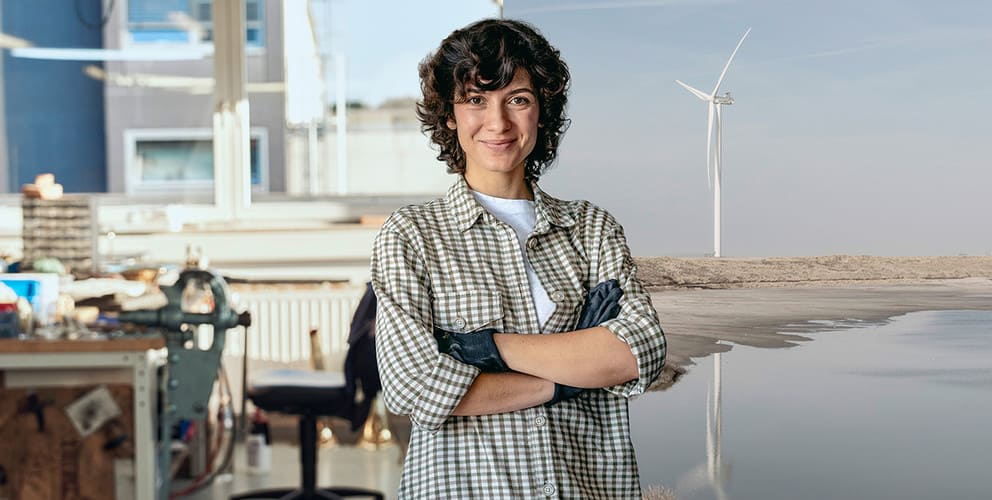 Handwerkerin steht im Vordergrund. Eine Werkstatt und eine Onshore Windkraftanlage sind im Hintergrund.
