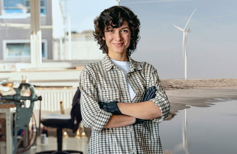 Handwerkerin steht im Vordergrund. Eine Werkstatt und eine Onshore Windkraftanlage sind im Hintergrund.