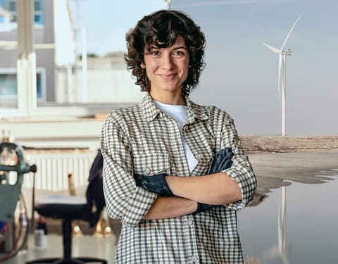 Handwerkerin steht im Vordergrund. Eine Werkstatt und eine Onshore Windkraftanlage sind im Hintergrund.
