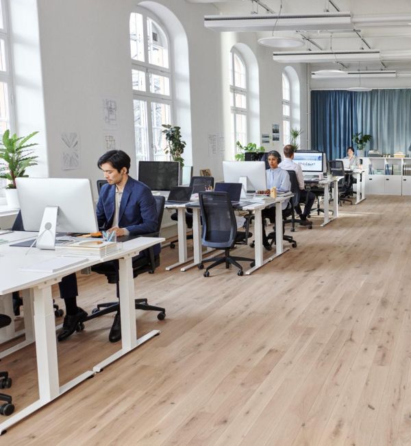 Mitarbeitende arbeiten in einem hellen, modernen Großraumbüro mit Holzboden und hohen Rundbogenfenstern.