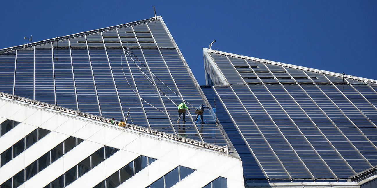 Zwei Arbeiter installieren Solarpaneele auf dem Dach eines modernen Gewerbegebäudes vor blauem Himmel.