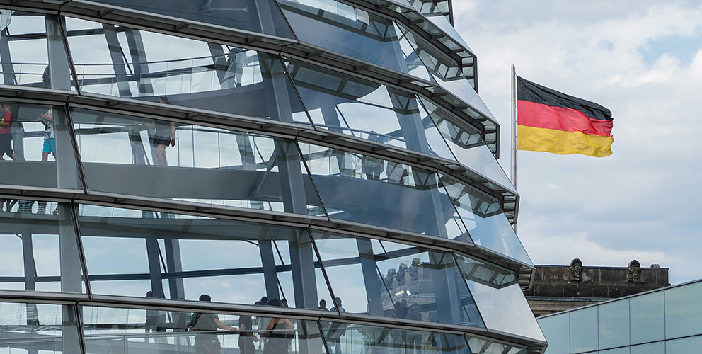 Ein Ausschnitt der Reichstagskuppel ist sichtbar. Dahinter weht eine Deutschlandfahne.