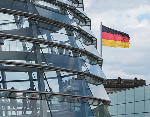 Ein Ausschnitt der Reichstagskuppel ist sichtbar. Dahinter weht eine Deutschlandfahne.