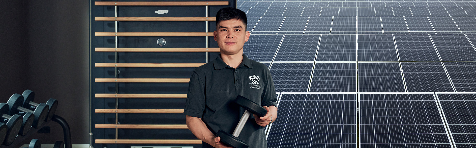 Ein Mann steht vor einem Hintergrund, der halb aus einem Gym und halb aus Solarmodulen besteht. Er hält eine Hantel in der Hand.