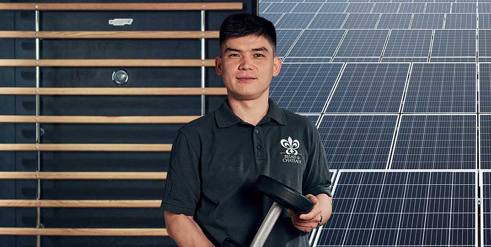 Ein Mann steht vor einem Hintergrund, der halb aus einem Gym und halb aus Solarmodulen besteht. Er hält eine Hantel in der Hand.