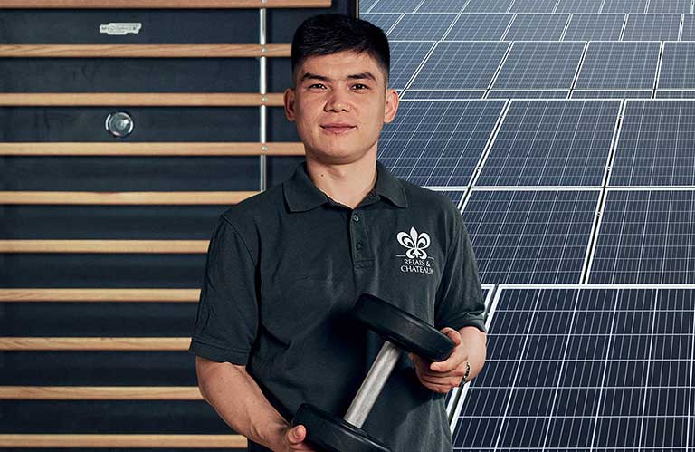 Ein Mann steht vor einem Hintergrund, der halb aus einem Gym und halb aus Solarmodulen besteht. Er hält eine Hantel in der Hand.