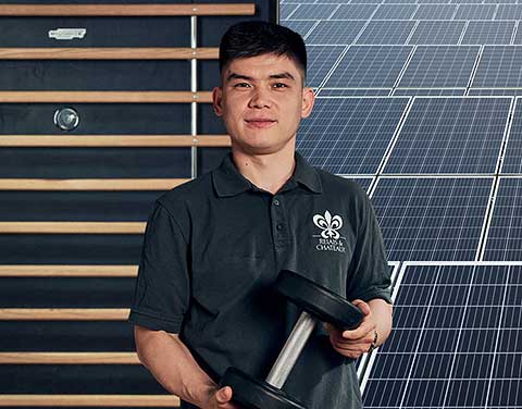 Ein Mann steht vor einem Hintergrund, der halb aus einem Gym und halb aus Solarmodulen besteht. Er hält eine Hantel in der Hand.