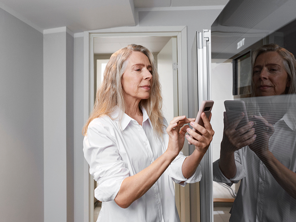 Das Bild zeigt eine ältere Frau, die in einem modernen Raum auf ihrem Smartphone tippt. Sie trägt ein weißes Hemd und schaut konzentriert auf ihr Handy. Im Hintergrund ist ein glänzender Kühlschrank zu sehen, und der Raum hat eine minimalistische, moderne Atmosphäre. Die Frau ist in einem neutralen Raum mit hellen Farben und einem modernen Design, was die sachliche und ruhige Stimmung des Bildes unterstreicht.