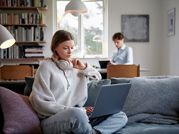 Frau sitzt auf einem Sofa und trinkt aus einer Tasse, blickt zur Kamera; im Hintergrund arbeitet eine Person am Laptop am Tisch – helles, gemütliches Wohnzimmer mit Regal, Lampe und Fenster.