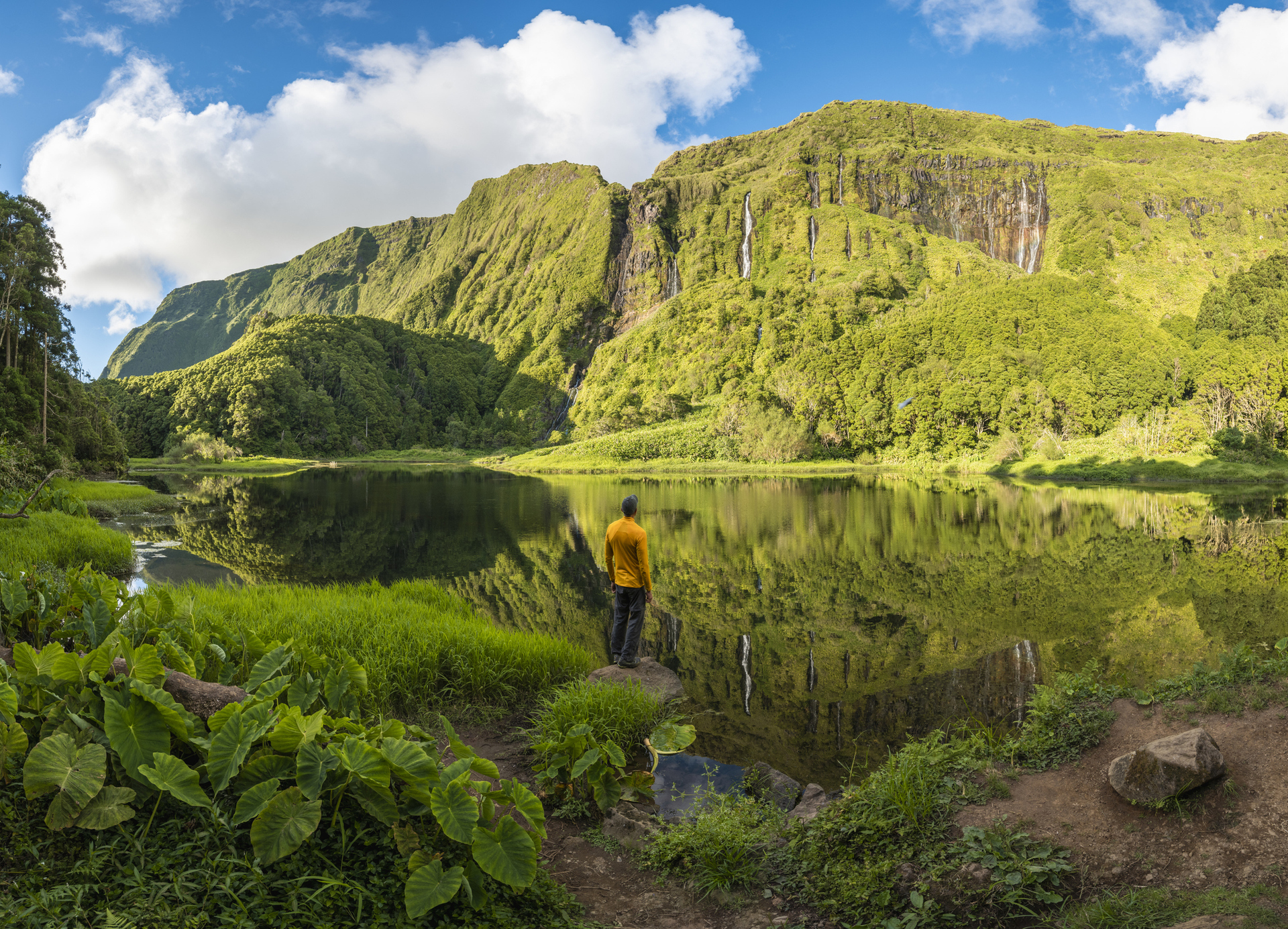 Person mit gelber Jacke blickt auf einen See vor grüner Bergkulisse
