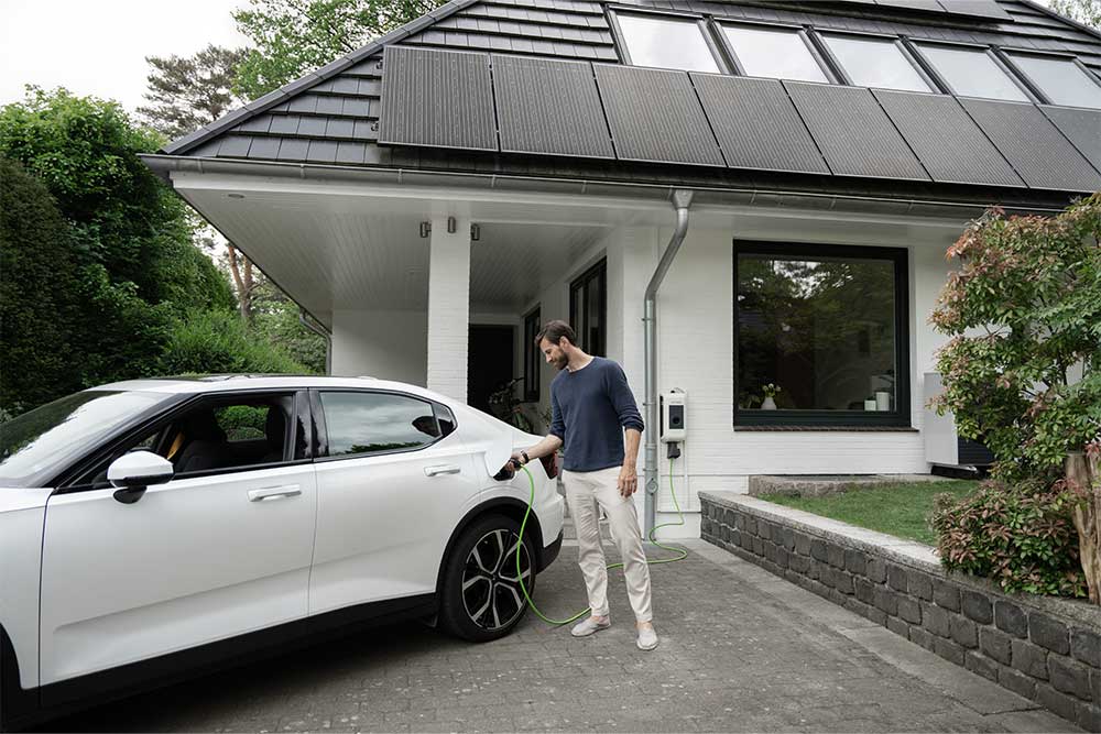 Mann steht am Elektroauto vor einem Haus mit PV-Anlage