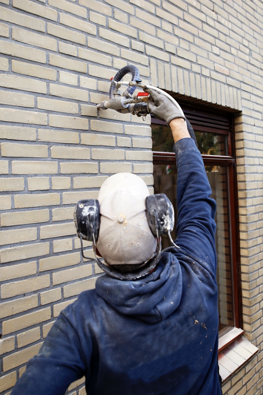 Ein Handwerker hält eine Einblasmaschine an ein Loch im Mauerwerk. So bringt er die Hohlraumdämmung nachträglich ein.