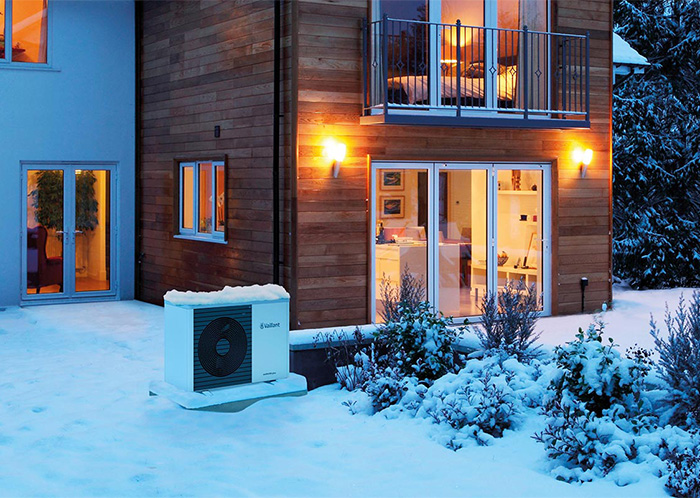 Eine Wärmepumpe steht vor einem beleuchteten Neubau aus Holz in einem verschneiten Garten