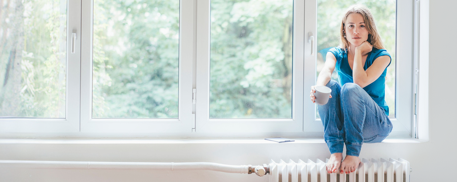 Frau sitzt mit Kaffeetasse auf der Fensterbank, mit den Füßen auf der Heizung.