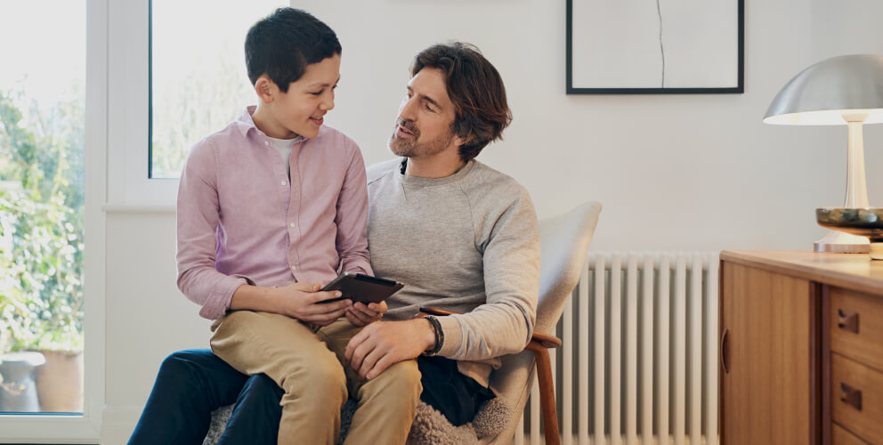 Ein Junge sitzt mit seinem Tablet in der Hand auf dem Schoß seines Vaters und erklärt ihm etwas. Der Vater hört gespannt zu. Beide befinden sich in einem hellen und modernen Raum.