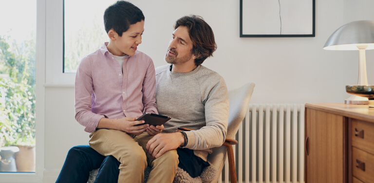 Ein Junge sitzt mit seinem Tablet in der Hand auf dem Schoß seines Vaters und erklärt ihm etwas. Der Vater hört gespannt zu. Beide befinden sich in einem hellen und modernen Raum.