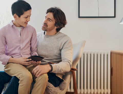 Ein Junge sitzt mit seinem Tablet in der Hand auf dem Schoß seines Vaters und erklärt ihm etwas. Der Vater hört gespannt zu. Beide befinden sich in einem hellen und modernen Raum.