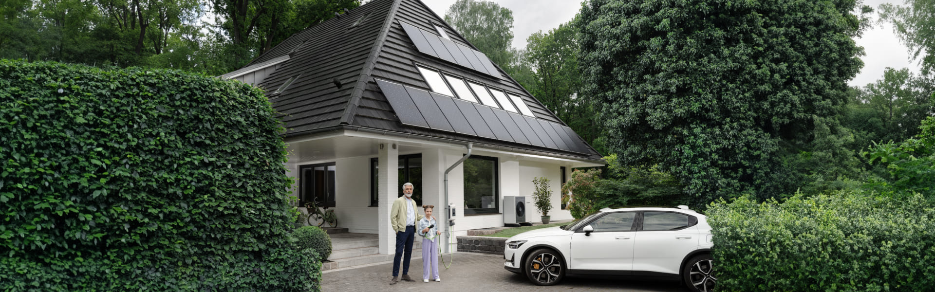 Ein Mann steht neben einem Kind vor einem Einfamilienhaus mit Solarpanelen auf dem Dach. Neben ihnen parkt ein Elektroauto in der Einfahrt.